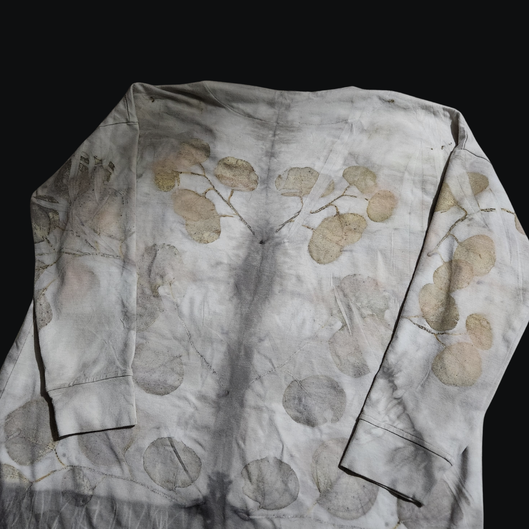 A close-up of a light-colored long-sleeved cardigan, adorned with a subtle pattern of eucalyptus leaves, created through a natural dye process. The shirt rests on a dark background, evoking the hushed stillness of a forest floor.